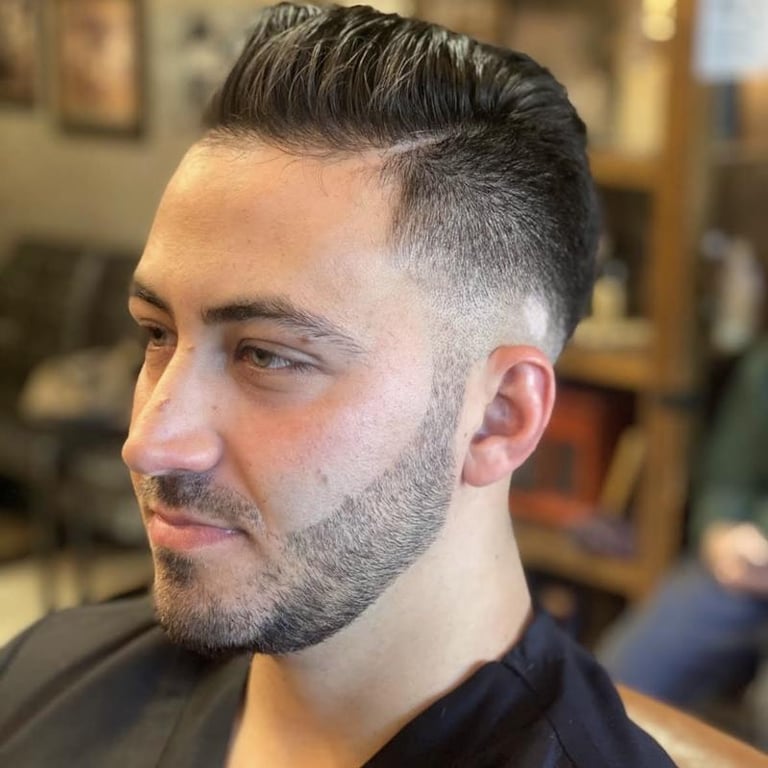 Man with styled dark hair in a pompadour cut and full beard, shown in profile view in a barbershop setting